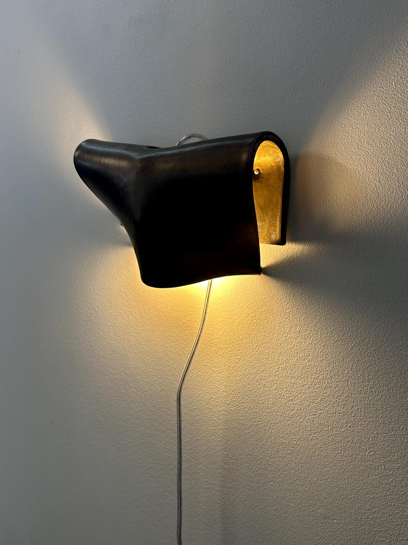 May include: A wall-mounted lamp featuring a curved, dark brown leather-like exterior shell that wraps around a metallic gold interior. The fixture emits a warm, glowing light from the top and bottom openings, connected by a thin, light-coloured power cord.