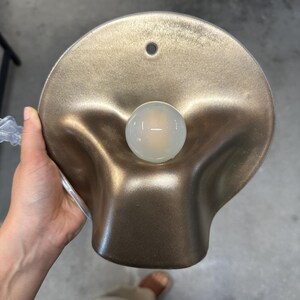 May include: A wall-mounted lamp featuring a metallic bronze, organic, undulating shade with a circular mounting hole at the top. A single, spherical, frosted white light bulb is centered within the recessed, curved basin of the fixture.