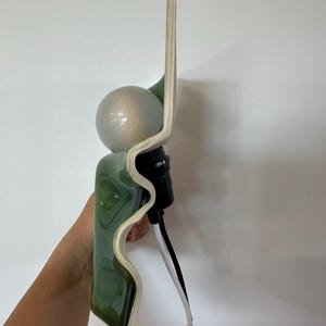 May include: A wall sconce featuring a wavy, glossy green ceramic backplate with a cream-colored edge. A spherical, frosted glass light bulb is mounted in the center, attached to a black socket with a white and black electrical cord extending downward.