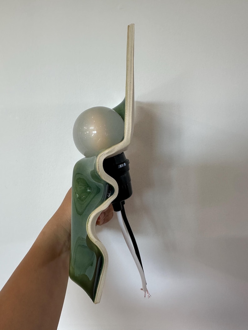 May include: A wall sconce featuring a wavy, glossy green ceramic backplate with a cream-colored edge. A spherical, frosted glass light bulb is mounted in the center, attached to a black socket with a white and black electrical cord extending downward.