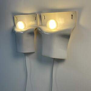 May include: A pair of white ceramic wall sconces with a smooth, curved, sculptural form. Each fixture features a single glowing round light bulb positioned in a recessed top section, with a flat back plate and a white power cord extending downward.