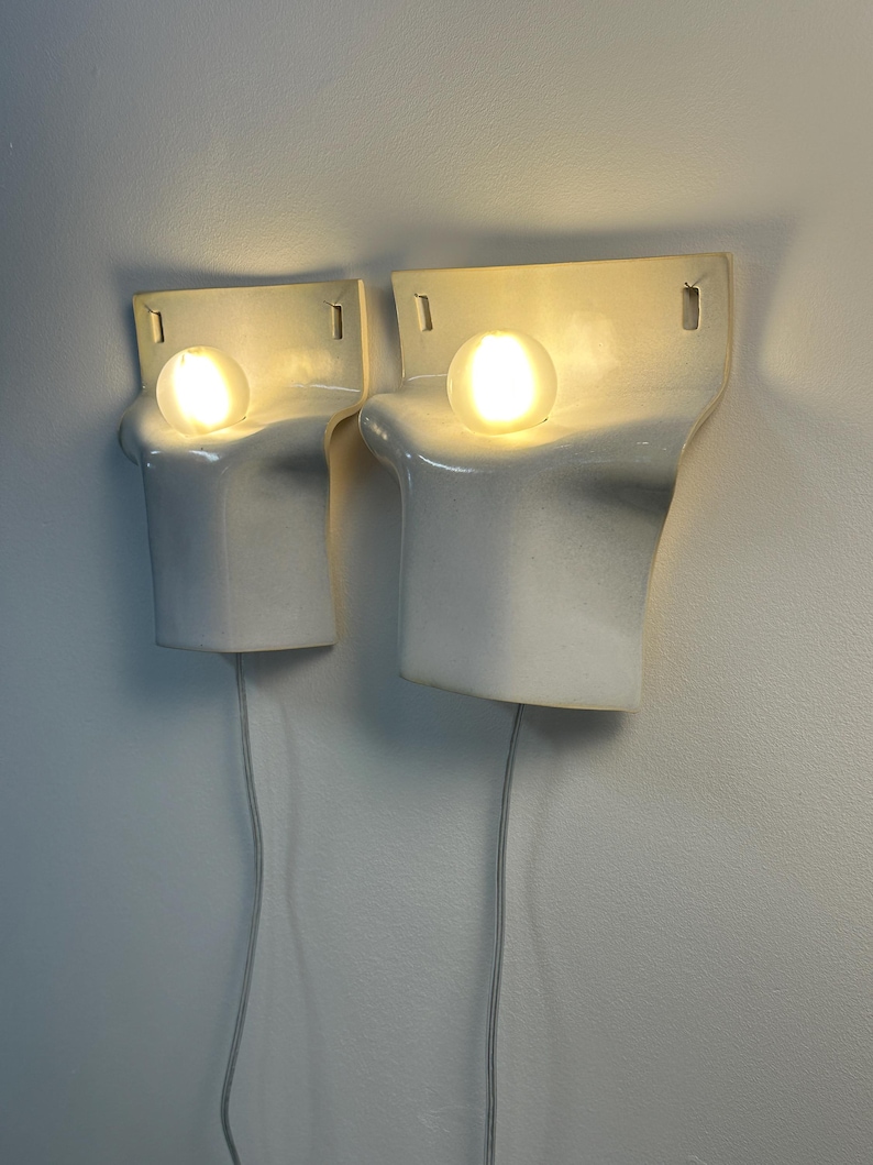 May include: A pair of white ceramic wall sconces with a smooth, curved, sculptural form. Each fixture features a single glowing round light bulb positioned in a recessed top section, with a flat back plate and a white power cord extending downward.