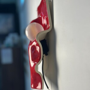 May include: A wall-mounted light fixture featuring a glossy red, wavy ceramic or plastic panel with a white, spherical light bulb protruding from the center. The fixture includes a black socket and a black electrical cord extending downward.