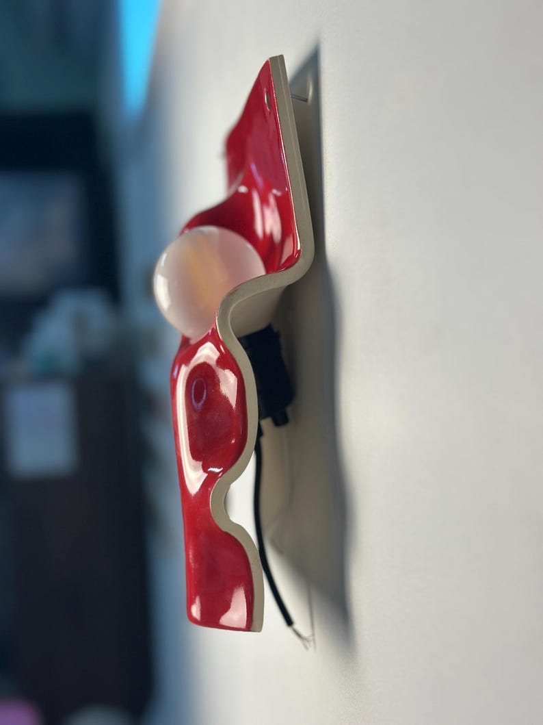 May include: A wall-mounted light fixture featuring a glossy red, wavy ceramic or plastic panel with a white, spherical light bulb protruding from the center. The fixture includes a black socket and a black electrical cord extending downward.