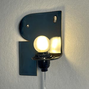 May include: A wall-mounted lamp featuring a glossy, dark blue ceramic backplate with an irregular, curved shape. A black socket holds a glowing, spherical white light bulb, and a thin, clear electrical cord extends downwards from the base.