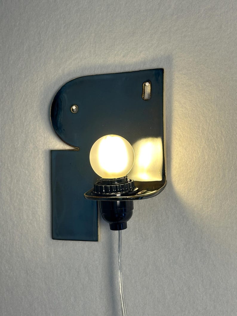 May include: A wall-mounted lamp featuring a glossy, dark blue ceramic backplate with an irregular, curved shape. A black socket holds a glowing, spherical white light bulb, and a thin, clear electrical cord extends downwards from the base.
