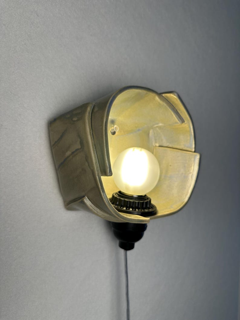 Puede incluir: Un aplique de pared con una pantalla de cer&aacute;mica de forma irregular hecha a mano y acabado amarillo p&aacute;lido brillante. La pantalla alberga una bombilla redonda de luz blanca esmerilada en un casquillo negro, con un cable fino hacia abajo.