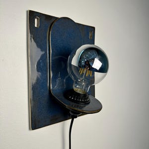 May include: A wall-mounted ceramic lamp featuring a glossy, deep blue rectangular backplate with rounded corners and a matching circular protrusion. A black socket holds a clear, spherical light bulb with a reflective silver-toned top half.