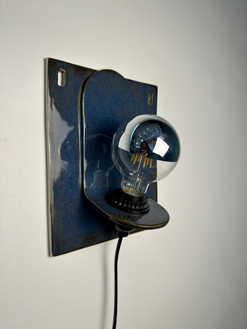 May include: A wall-mounted ceramic lamp featuring a glossy, deep blue rectangular backplate with rounded corners and a matching circular protrusion. A black socket holds a clear, spherical light bulb with a reflective silver-toned top half.