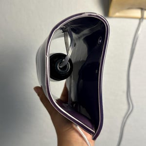 May include: A curved, deep purple wall lamp fixture with a glossy finish. The interior features a black circular light socket housing and a clear electrical cord that extends from the base, designed for mounting against a flat vertical surface.