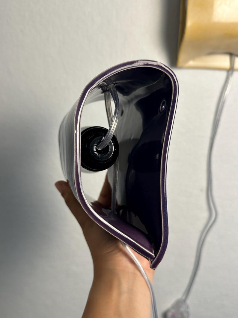 May include: A curved, deep purple wall lamp fixture with a glossy finish. The interior features a black circular light socket housing and a clear electrical cord that extends from the base, designed for mounting against a flat vertical surface.