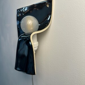 K&ouml;nnte beinhalten: Eine Wandleuchte mit einem gl&auml;nzend schwarzen Keramikschirm in geschwungenem, gefaltetem Design. Die Leuchte verf&uuml;gt &uuml;ber eine wei&szlig;e Lampenfassung und eine runde, mattwei&szlig;e Gl&uuml;hbirne. Ein d&uuml;nnes, transparentes Stromkabel f&uuml;hrt nach unten.