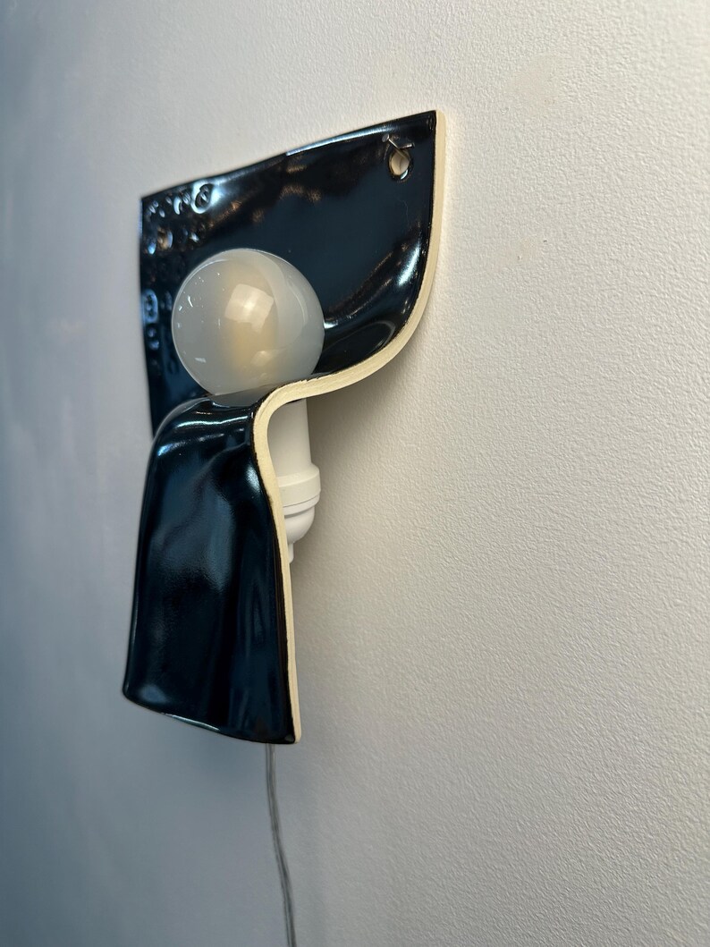 K&ouml;nnte beinhalten: Eine Wandleuchte mit einem gl&auml;nzend schwarzen Keramikschirm in geschwungenem, gefaltetem Design. Die Leuchte verf&uuml;gt &uuml;ber eine wei&szlig;e Lampenfassung und eine runde, mattwei&szlig;e Gl&uuml;hbirne. Ein d&uuml;nnes, transparentes Stromkabel f&uuml;hrt nach unten.