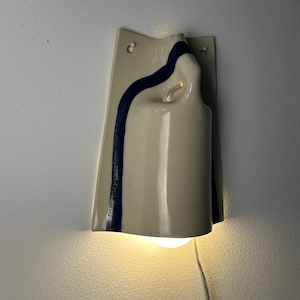May include: A wall-mounted ceramic sconce featuring a glossy cream-colored finish with a single, vertical dark blue stripe. The fixture has a curved, sculptural form with two mounting holes at the top and a light bulb emitting a warm glow from below.