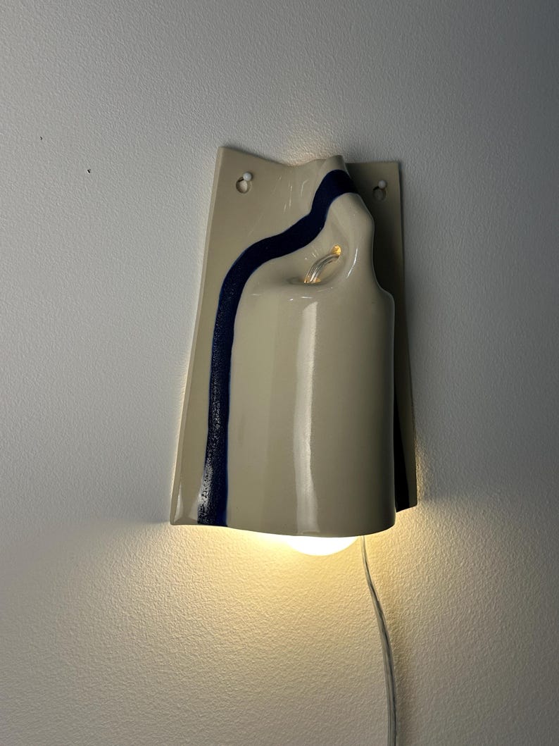 May include: A wall-mounted ceramic sconce featuring a glossy cream-colored finish with a single, vertical dark blue stripe. The fixture has a curved, sculptural form with two mounting holes at the top and a light bulb emitting a warm glow from below.