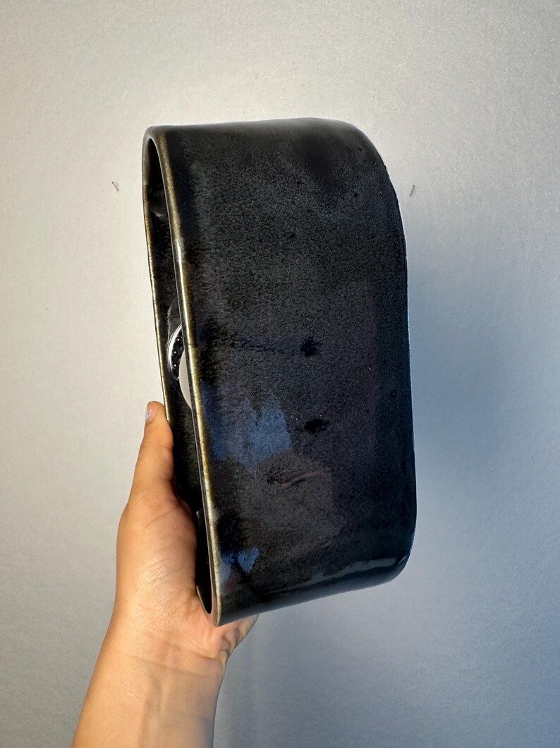 May include: A black ceramic wall-mounted soap dish with a curved design. The dish has a glossy finish with a dark bronze trim around the edges. The soap dish is held in a hand.