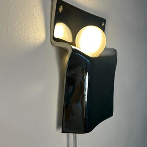 Peut inclure: Une applique murale compos&eacute;e d'un corps en c&eacute;ramique noire brillante au design incurv&eacute; et pli&eacute;. Une ampoule sph&eacute;rique en verre d&eacute;poli blanc est log&eacute;e dans la courbe sup&eacute;rieure, diffusant une lumi&egrave;re chaude. Un fin cordon &eacute;lectrique transparent pend.