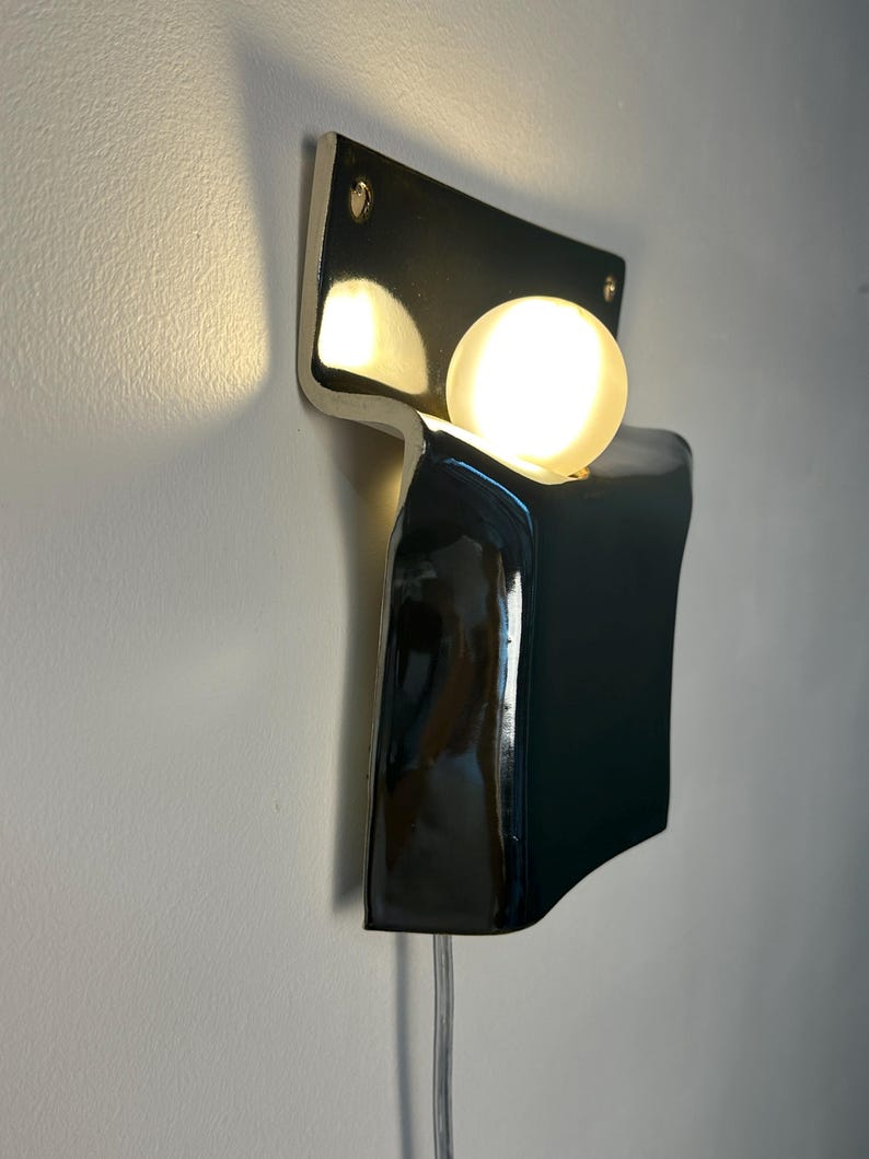 Peut inclure: Une applique murale compos&eacute;e d'un corps en c&eacute;ramique noire brillante au design incurv&eacute; et pli&eacute;. Une ampoule sph&eacute;rique en verre d&eacute;poli blanc est log&eacute;e dans la courbe sup&eacute;rieure, diffusant une lumi&egrave;re chaude. Un fin cordon &eacute;lectrique transparent pend.