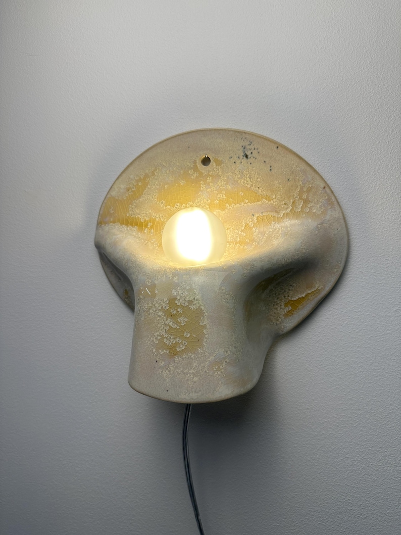 May include: A wall-mounted ceramic sconce featuring a rounded, organic shape with a mottled cream and yellow glaze. A single glowing light bulb is recessed into the center of the fixture, which includes a small hole at the top for mounting.