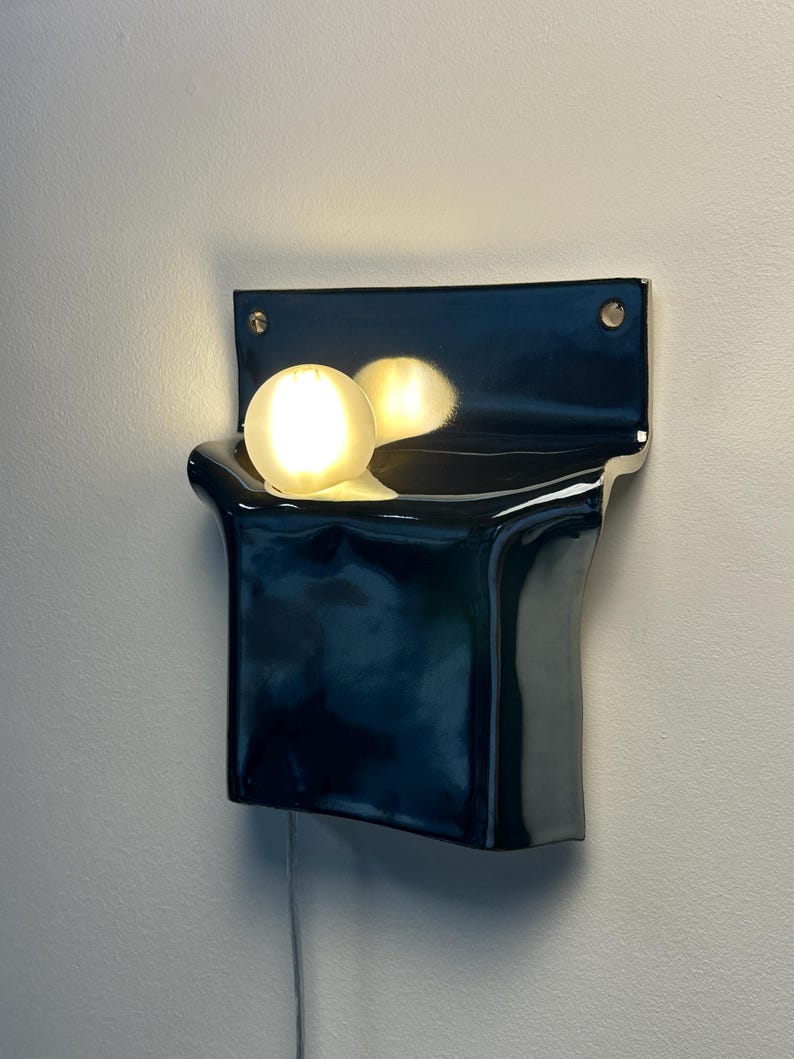 Peut inclure: Une applique murale en c&eacute;ramique noire avec une finition brillante et une plaque arri&egrave;re rectangulaire dot&eacute;e de deux trous de fixation. Le luminaire comprend une ampoule blanche sph&eacute;rique qui &eacute;met une lueur chaude et se refl&egrave;te sur la surface.