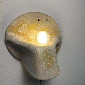 May include: A wall-mounted ceramic sconce featuring a curved, organic shape with a textured, mottled yellow and off-white glaze. A single, glowing, spherical frosted glass bulb is nestled within the center of the fixture, emitting a warm light.