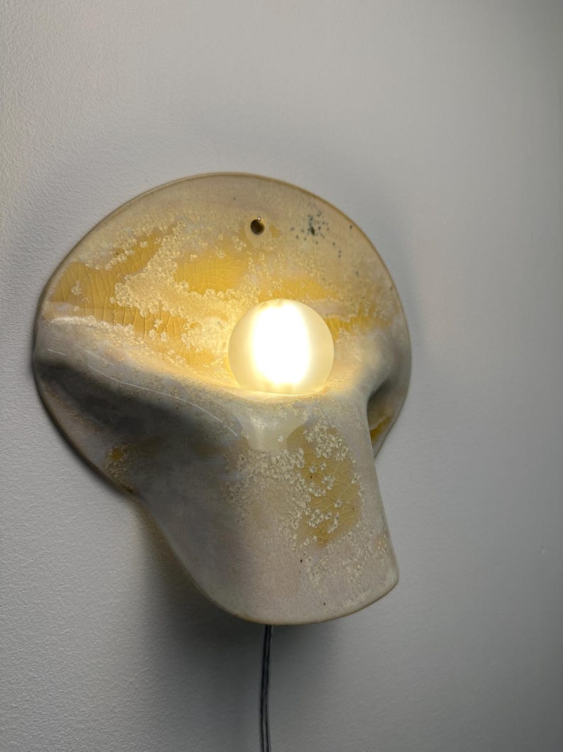 May include: A wall-mounted ceramic sconce featuring a curved, organic shape with a textured, mottled yellow and off-white glaze. A single, glowing, spherical frosted glass bulb is nestled within the center of the fixture, emitting a warm light.