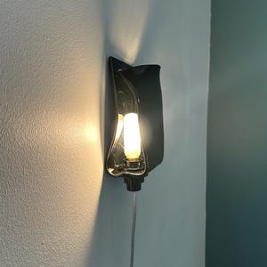 May include: A wall-mounted sconce featuring a glossy black, curved, wave-like ceramic shade. The fixture holds a single, vertically oriented, warm-toned light bulb that casts a soft glow against the wall. A clear power cord extends downward.