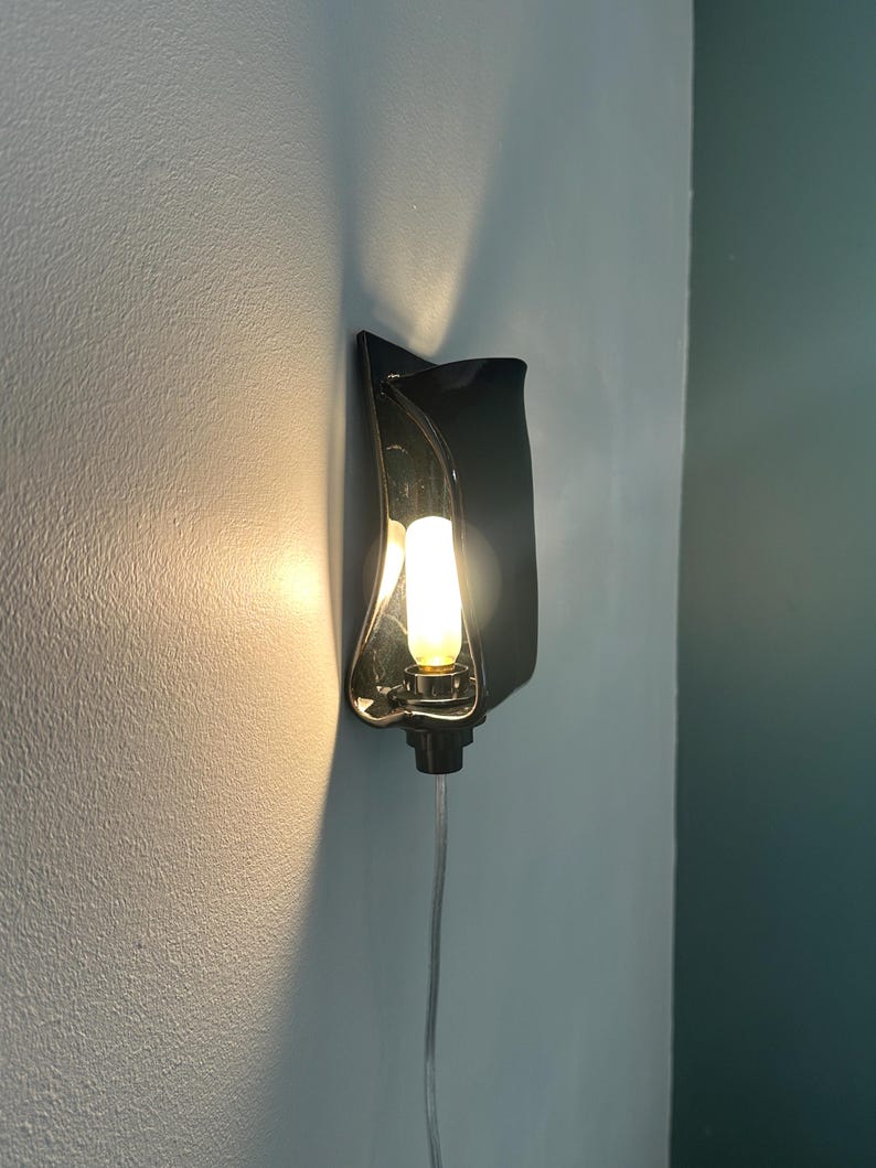 May include: A wall-mounted sconce featuring a glossy black, curved, wave-like ceramic shade. The fixture holds a single, vertically oriented, warm-toned light bulb that casts a soft glow against the wall. A clear power cord extends downward.