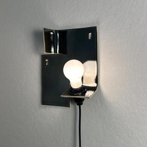 Pode incluir: Uma arandela de parede preta moderna com um design geom&eacute;trico curvo. A arandela apresenta uma &uacute;nica l&acirc;mpada redonda e iluminada. Um cabo preto estende-se para baixo a partir da lumin&aacute;ria.