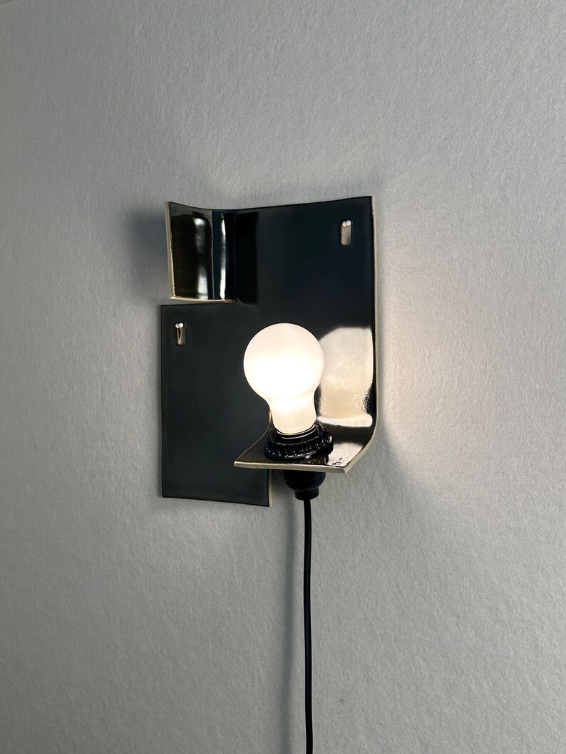 Pode incluir: Uma arandela de parede preta moderna com um design geom&eacute;trico curvo. A arandela apresenta uma &uacute;nica l&acirc;mpada redonda e iluminada. Um cabo preto estende-se para baixo a partir da lumin&aacute;ria.