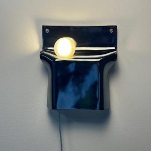 Peut inclure: Une applique murale compos&eacute;e d'une base en c&eacute;ramique noire brillante au design incurv&eacute; et pli&eacute;. Une ampoule sph&eacute;rique blanche givr&eacute;e est plac&eacute;e au centre de la courbe sup&eacute;rieure, diffusant une lumi&egrave;re chaude. Un fin cordon gris pend vers le bas.