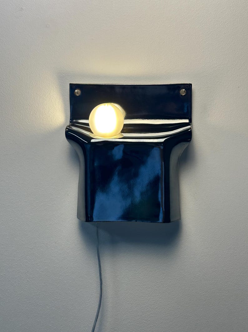 Peut inclure: Une applique murale compos&eacute;e d'une base en c&eacute;ramique noire brillante au design incurv&eacute; et pli&eacute;. Une ampoule sph&eacute;rique blanche givr&eacute;e est plac&eacute;e au centre de la courbe sup&eacute;rieure, diffusant une lumi&egrave;re chaude. Un fin cordon gris pend vers le bas.