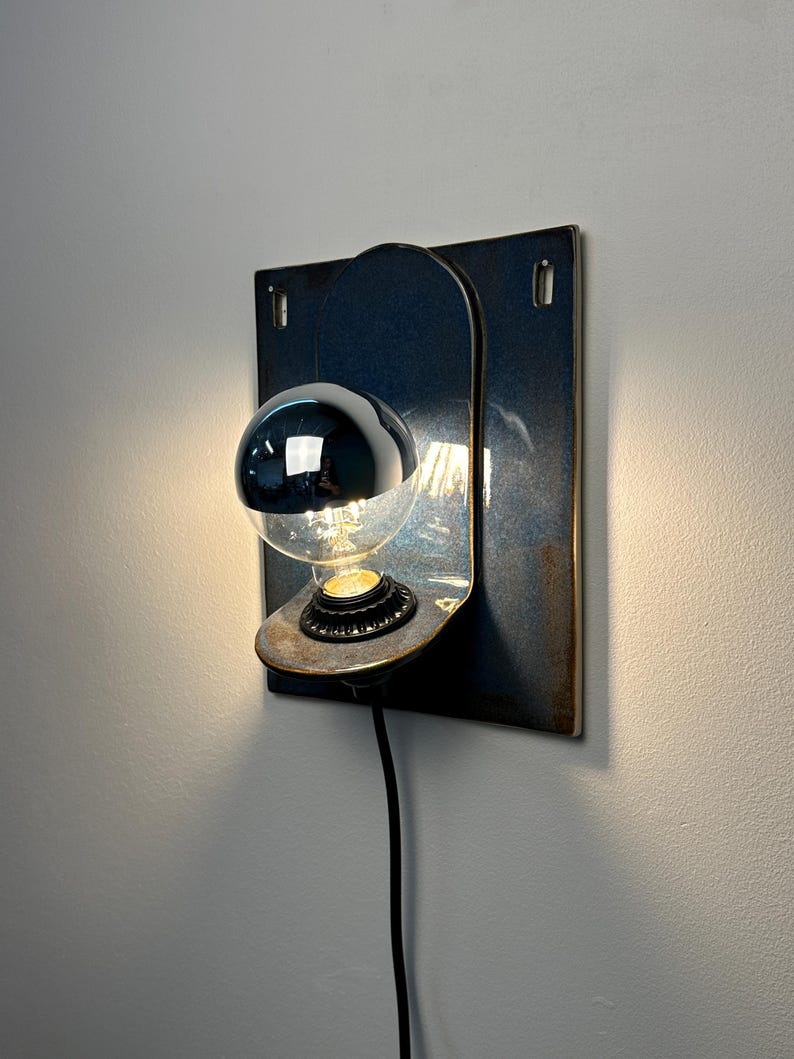May include: A wall-mounted lamp featuring a rectangular, dark blue ceramic backplate with a curved shelf holding a light bulb. The bulb has a reflective silver top half and a clear bottom half, emitting a warm glow from a black socket base.