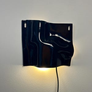 Puede incluir: Una l&aacute;mpara de pared con una pantalla de cer&aacute;mica azul marino brillante y superficie ondulada. La pantalla rectangular tiene dos ranuras verticales en la parte superior para el montaje y emite una luz c&aacute;lida hacia abajo desde un cable negro.