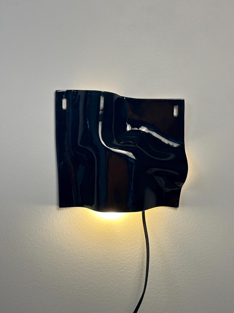 Puede incluir: Una l&aacute;mpara de pared con una pantalla de cer&aacute;mica azul marino brillante y superficie ondulada. La pantalla rectangular tiene dos ranuras verticales en la parte superior para el montaje y emite una luz c&aacute;lida hacia abajo desde un cable negro.