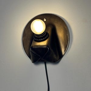May include: A wall-mounted lamp featuring a circular dark metal backplate with a protruding, folded form. A single round, frosted light bulb is mounted on the upper section of the plate, emitting a warm glow. A black power cord extends downward.