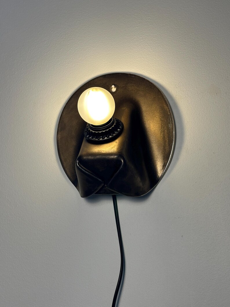 May include: A wall-mounted lamp featuring a circular dark metal backplate with a protruding, folded form. A single round, frosted light bulb is mounted on the upper section of the plate, emitting a warm glow. A black power cord extends downward.