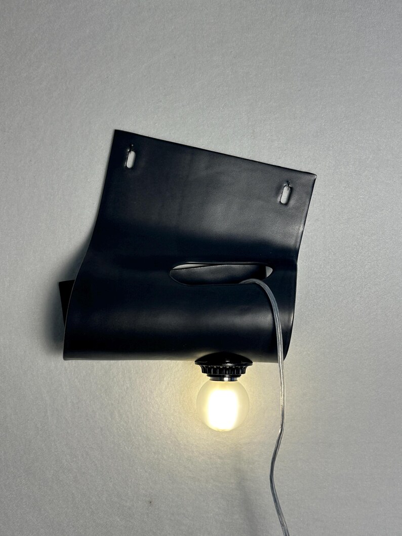 Puede incluir: Una l&aacute;mpara de pared negra que presenta una l&aacute;mina rectangular curvada con dos ranuras de montaje superiores y un recorte horizontal central. Una bombilla redonda esmerilada est&aacute; unida al borde inferior, conectada por un cable el&eacute;ctrico fino.