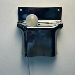 Peut inclure: Une applique murale compos&eacute;e d'une base en c&eacute;ramique noire brillante aux courbes ondul&eacute;es. Une ampoule sph&eacute;rique en verre blanc d&eacute;poli est log&eacute;e dans l'indentation sup&eacute;rieure de la structure. Un fin cordon &eacute;lectrique de couleur claire pend vers le bas.