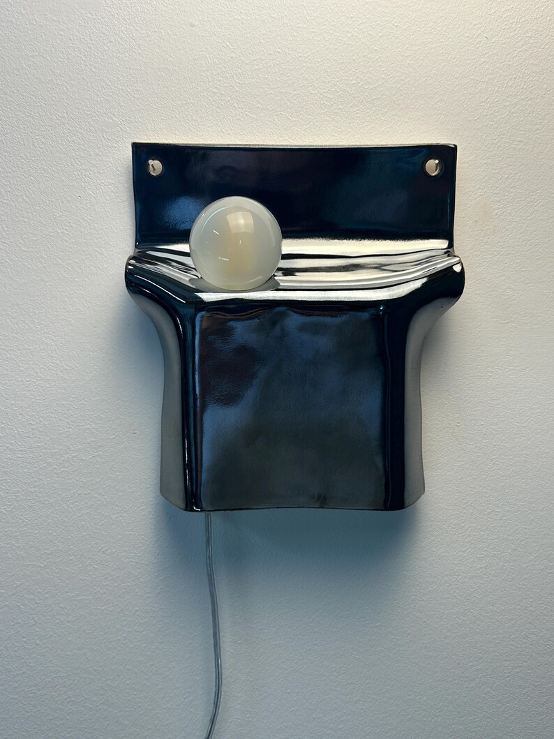 Peut inclure: Une applique murale compos&eacute;e d'une base en c&eacute;ramique noire brillante aux courbes ondul&eacute;es. Une ampoule sph&eacute;rique en verre blanc d&eacute;poli est log&eacute;e dans l'indentation sup&eacute;rieure de la structure. Un fin cordon &eacute;lectrique de couleur claire pend vers le bas.