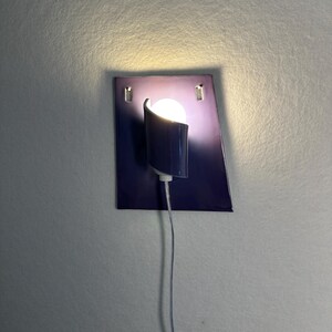 Pode incluir: Uma arandela de parede moderna com uma placa traseira retangular azul escuro e uma sombra curva e cil&iacute;ndrica. A arandela apresenta uma l&acirc;mpada redonda e brilhante e um cabo de alimenta&ccedil;&atilde;o branco fino. A lumin&aacute;ria &eacute; montada em uma parede cinza claro.