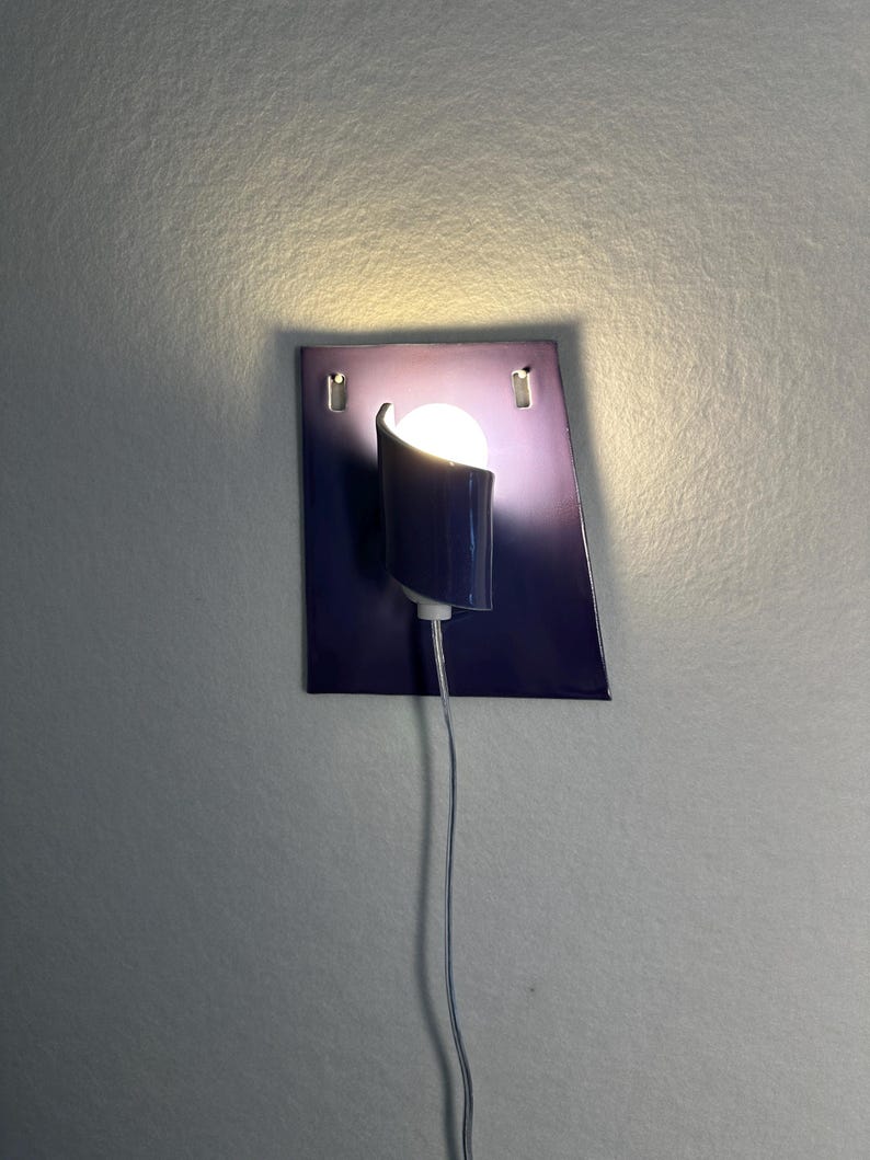 Pode incluir: Uma arandela de parede moderna com uma placa traseira retangular azul escuro e uma sombra curva e cil&iacute;ndrica. A arandela apresenta uma l&acirc;mpada redonda e brilhante e um cabo de alimenta&ccedil;&atilde;o branco fino. A lumin&aacute;ria &eacute; montada em uma parede cinza claro.