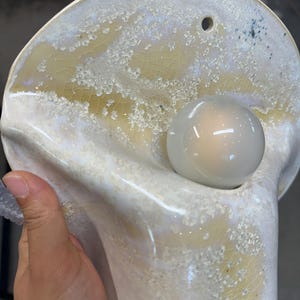 May include: A wall-mounted ceramic light fixture with a glossy, off-white and pale yellow crackle glaze finish. The organic, shell-like shape features a circular opening at the top for mounting and a central socket holding a round, frosted light bulb.