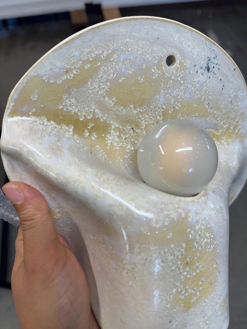 May include: A wall-mounted ceramic light fixture with a glossy, off-white and pale yellow crackle glaze finish. The organic, shell-like shape features a circular opening at the top for mounting and a central socket holding a round, frosted light bulb.