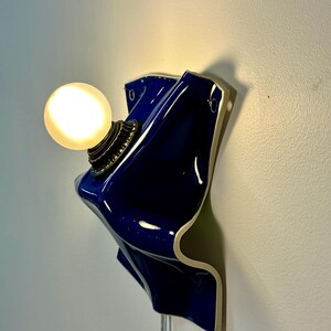 May include: A wall-mounted lamp featuring a glossy, deep blue, wavy ceramic base with a white edge. A round, frosted glass light bulb is attached to the center of the ceramic form by a dark, textured metal socket, emitting a warm, soft glow.