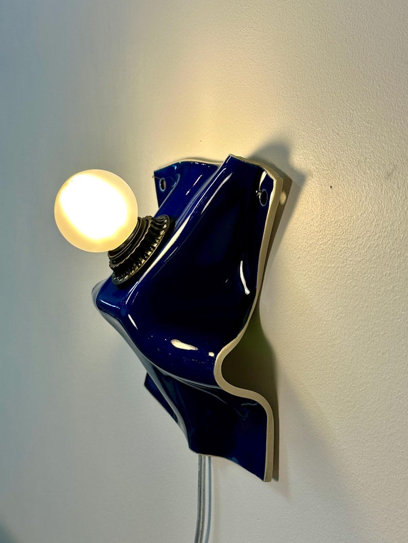 May include: A wall-mounted lamp featuring a glossy, deep blue, wavy ceramic base with a white edge. A round, frosted glass light bulb is attached to the center of the ceramic form by a dark, textured metal socket, emitting a warm, soft glow.