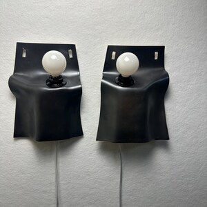 May include: A pair of wall-mounted lamps featuring matte black, curved, flexible-looking rectangular shades. Each lamp has a single white spherical light bulb centered in the upper portion of the shade and a thin light-colored power cord extending downward.