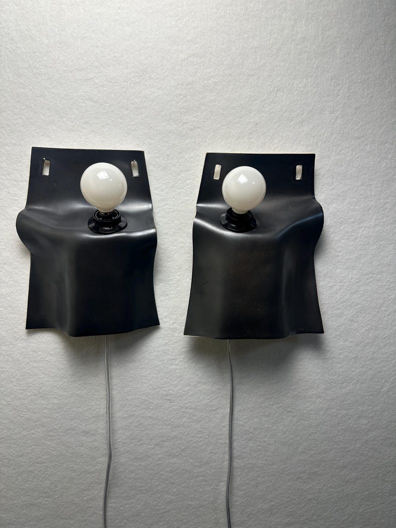 May include: A pair of wall-mounted lamps featuring matte black, curved, flexible-looking rectangular shades. Each lamp has a single white spherical light bulb centered in the upper portion of the shade and a thin light-colored power cord extending downward.