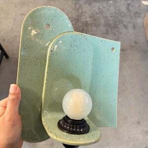 May include: A wall-mounted light fixture featuring a mint green ceramic backplate with dark speckles. The fixture has a rounded, arched shape with two pre-drilled holes for mounting and a black socket holding a round, frosted white light bulb.