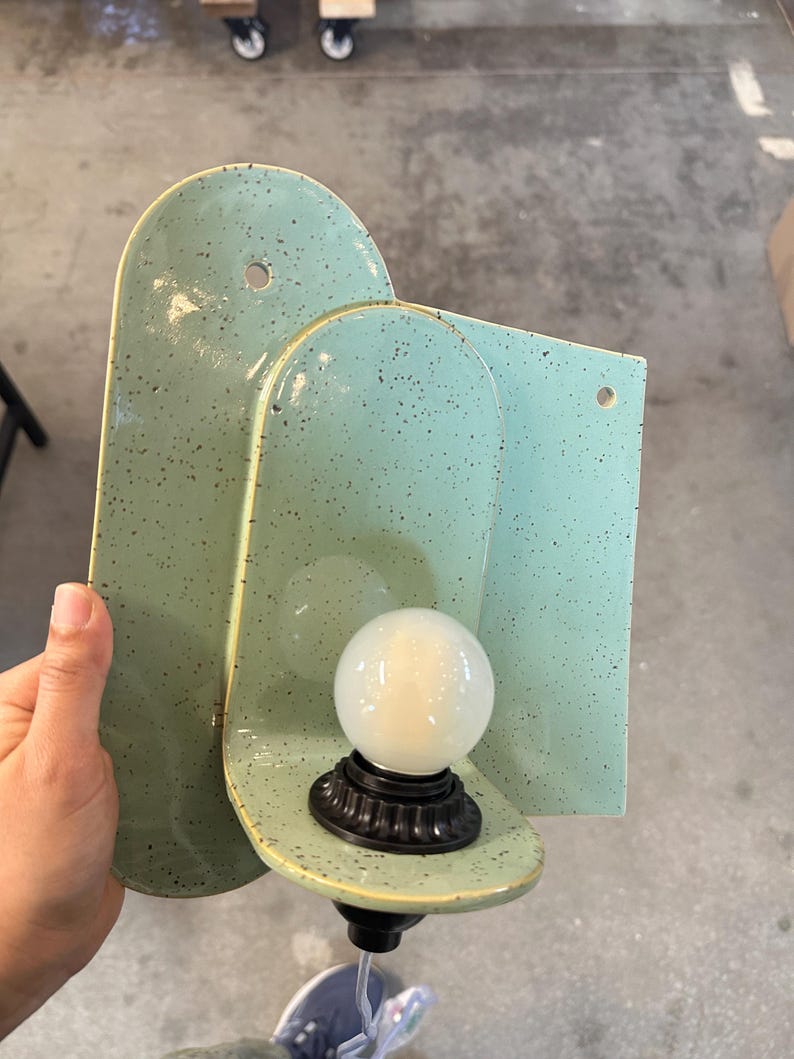 May include: A wall-mounted light fixture featuring a mint green ceramic backplate with dark speckles. The fixture has a rounded, arched shape with two pre-drilled holes for mounting and a black socket holding a round, frosted white light bulb.